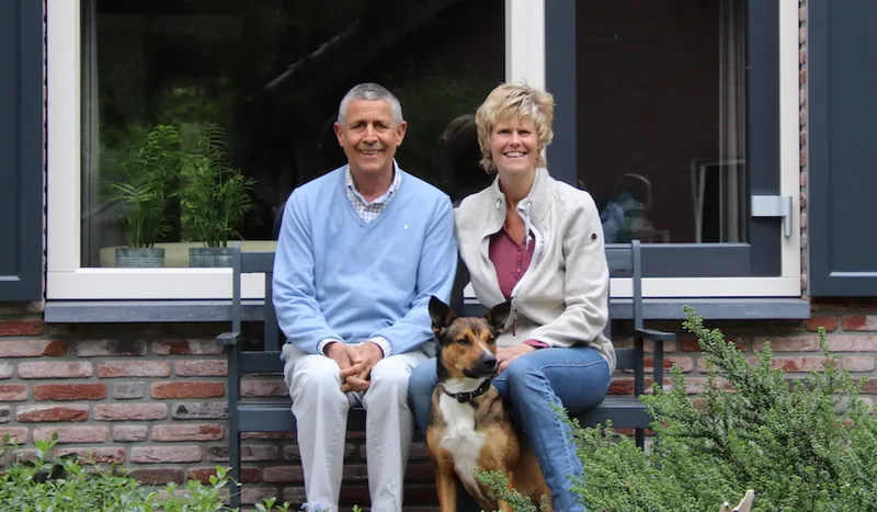 Een ouder stel met hun hond op een bankje voor hun huis