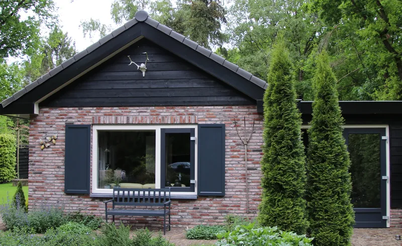 Een bakstenen bungalow met donkere houten gevel, luiken en een bankje, tussen hoge coniferen