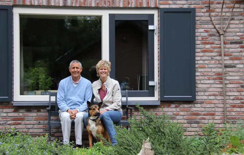 Een ouder stel met hun hond op een bankje voor hun bakstenen huis