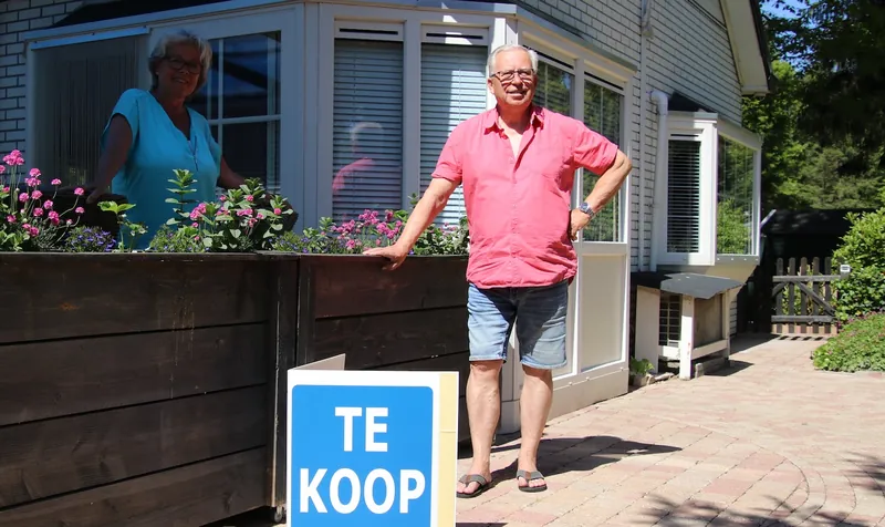 Een ouder stel bij hun huis, met een bord Te koop in de tuin