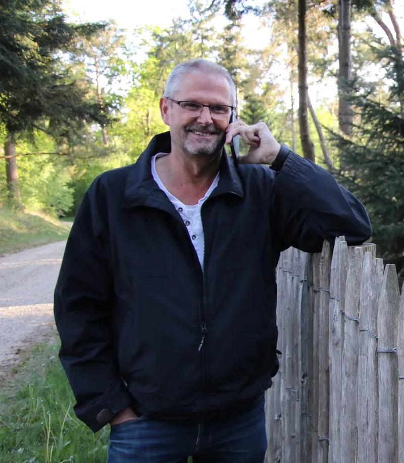 Joep van der Pol, lachend aan de telefoon, leunend op een houten hek aan de rand van het bos