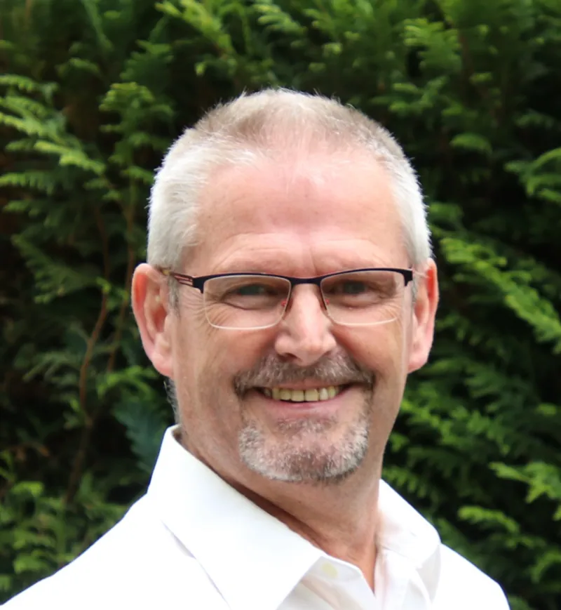 Joep van der Pol, lachend, in een wit overhemd voor een groene haag