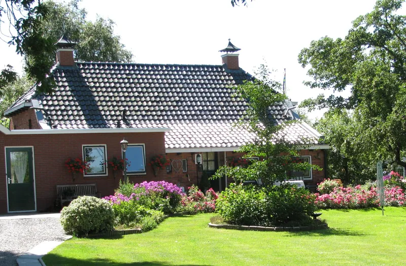 Een boerderijwoning met een rode pannenkap en een tuin vol bloeiende rozen en hortensia’s